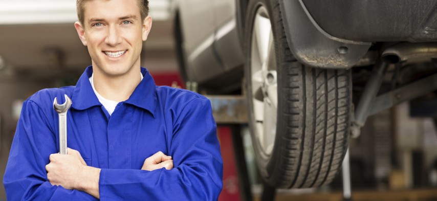Portrait of a happy young car mechanic holding wrench with car on hoist in background. Horizontal shot.
